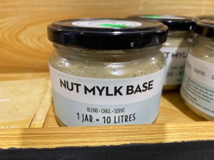 A shelf with jars that show the label 'Nut Mylk Base: blend, chill serve — 1 jar = 10 litres'